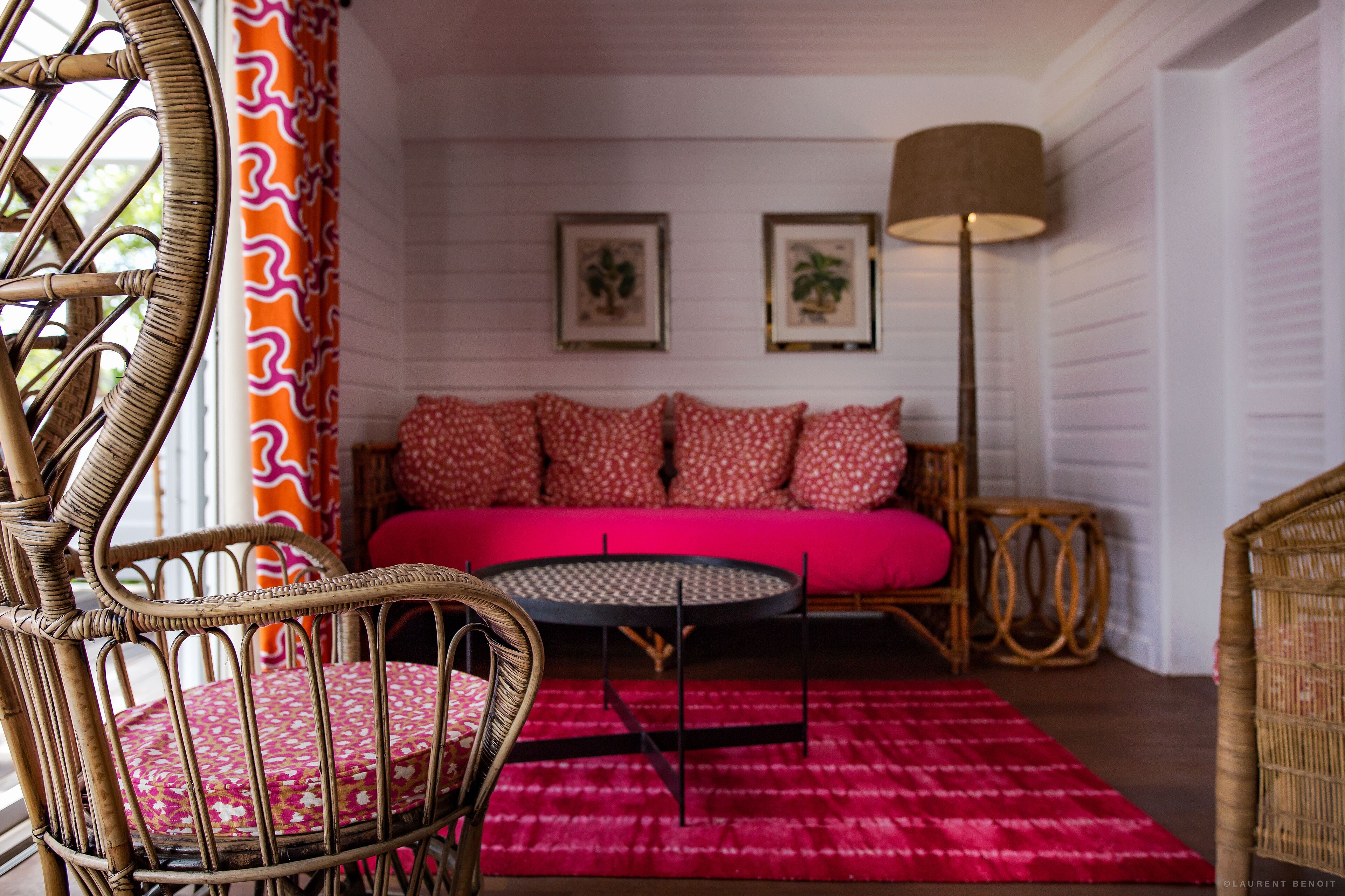 A wicker chair with a floral cushion, a round table, a sofa with red cushions, and a floor lamp.