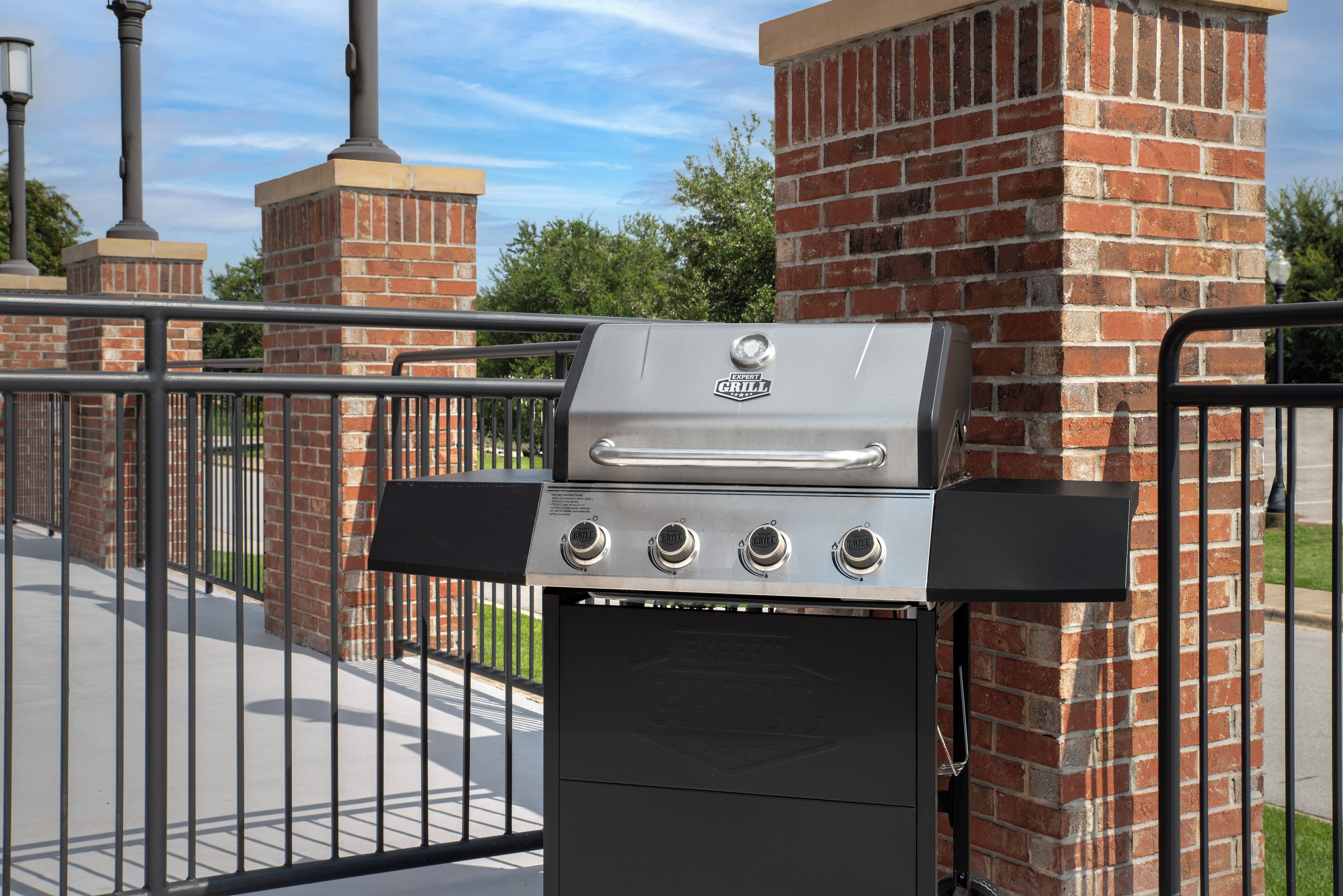 A barbecue grill with four burners, a control panel, and a chimney, set against a brick wall and metal fence.