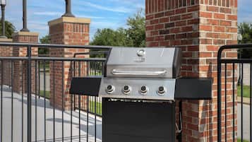 Un barbecue doté de quatre brûleurs, d’un panneau de commande et d’une cheminée, installé devant un mur de briques et une clôture en métal.