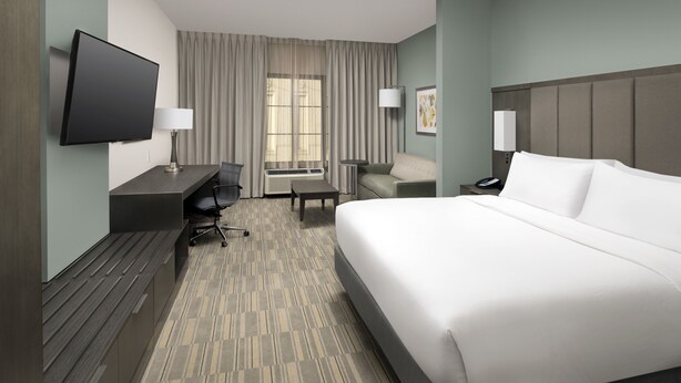 Quarto de hotel com uma cama grande, uma escrivaninha com cadeira, televisão de tela plana, abajur e janela com cortinas.