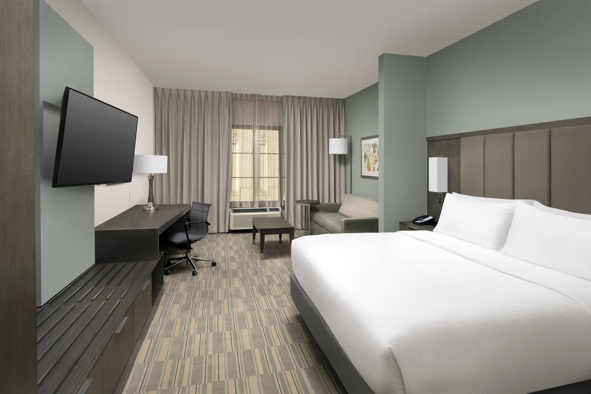 A hotel room with a large bed, a desk with a chair, a flat-screen TV, a lamp, and a window with curtains.