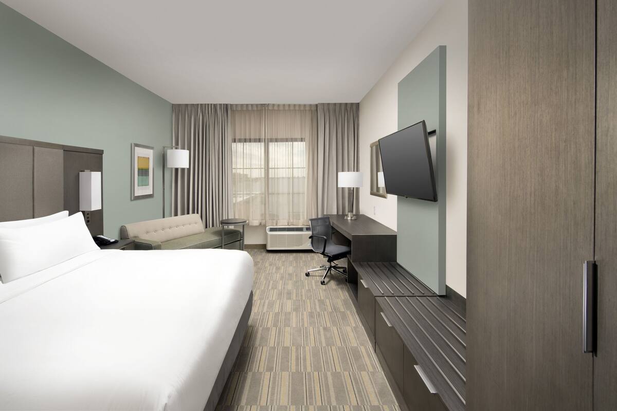 A hotel room with a large bed, a desk, a chair, a flat-screen TV, and a large window with curtains.
