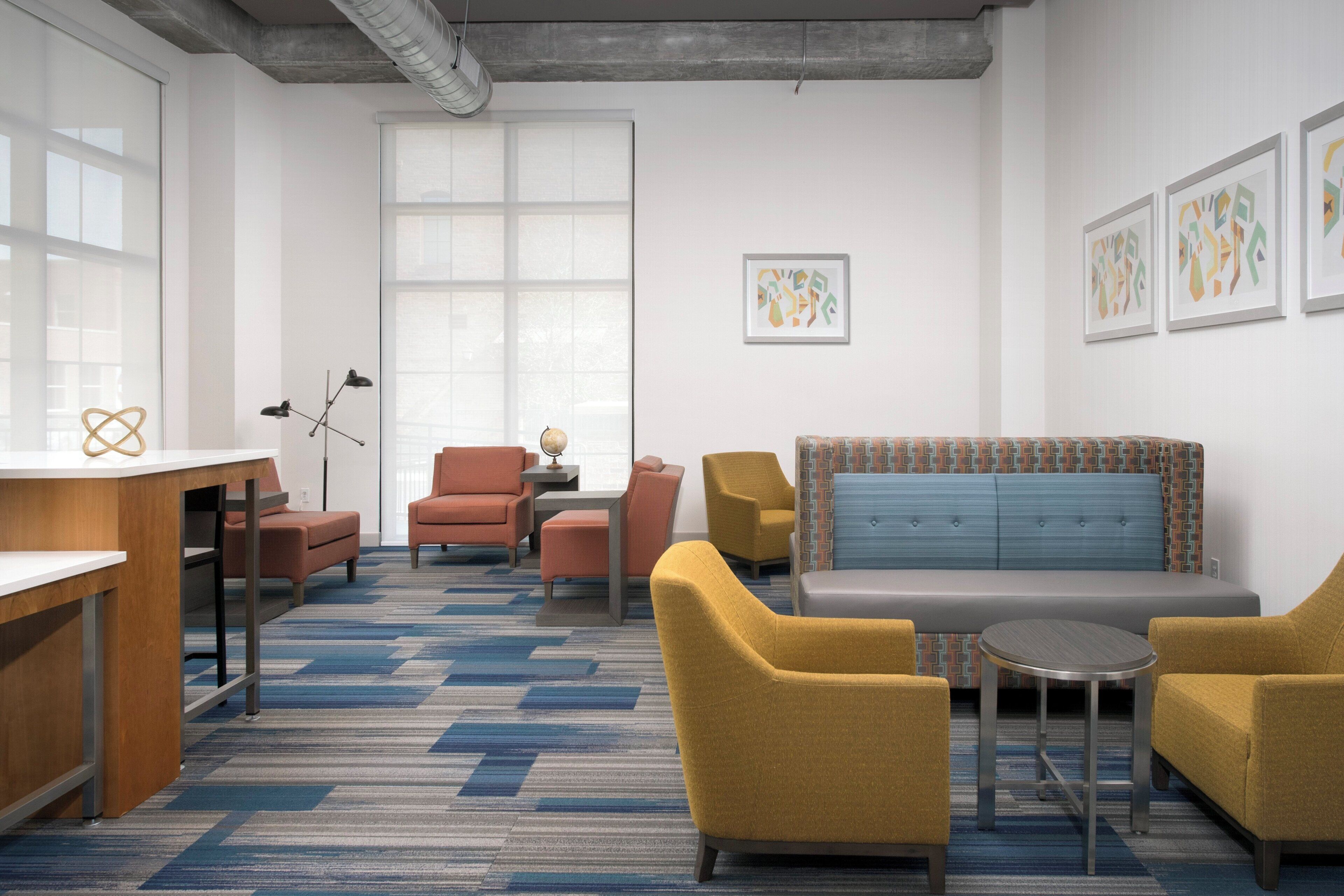 Ein moderner Lounge-Bereich mit einer Couch, Sesseln, einem kleinen Tisch und einem Schreibtisch. Es gibt Fenster mit Jalousien, einen Deckenlüfter und gerahmte Kunstwerke an der Wand.