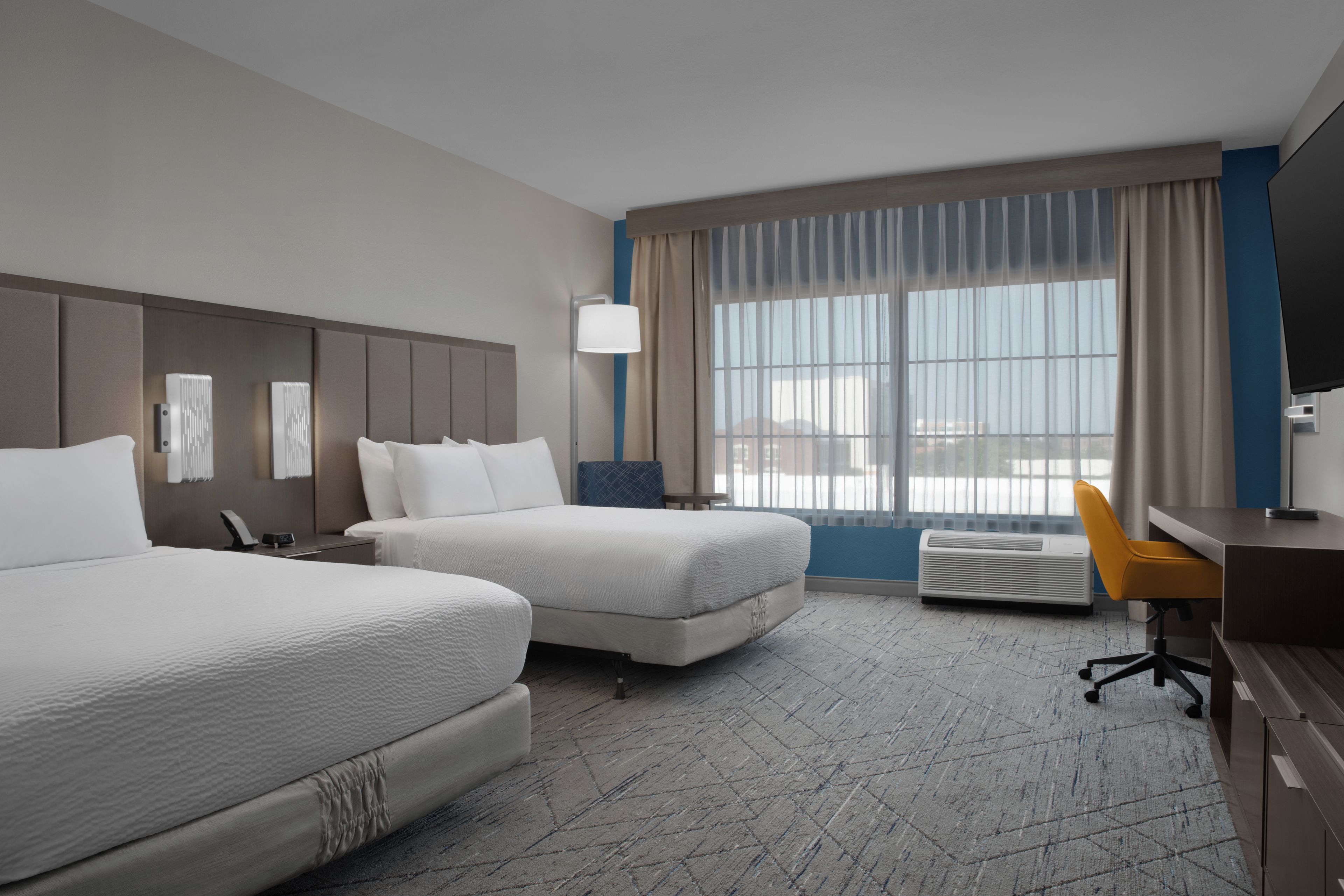 A hotel room with two beds, a desk, a chair, a lamp, and a large window with curtains.