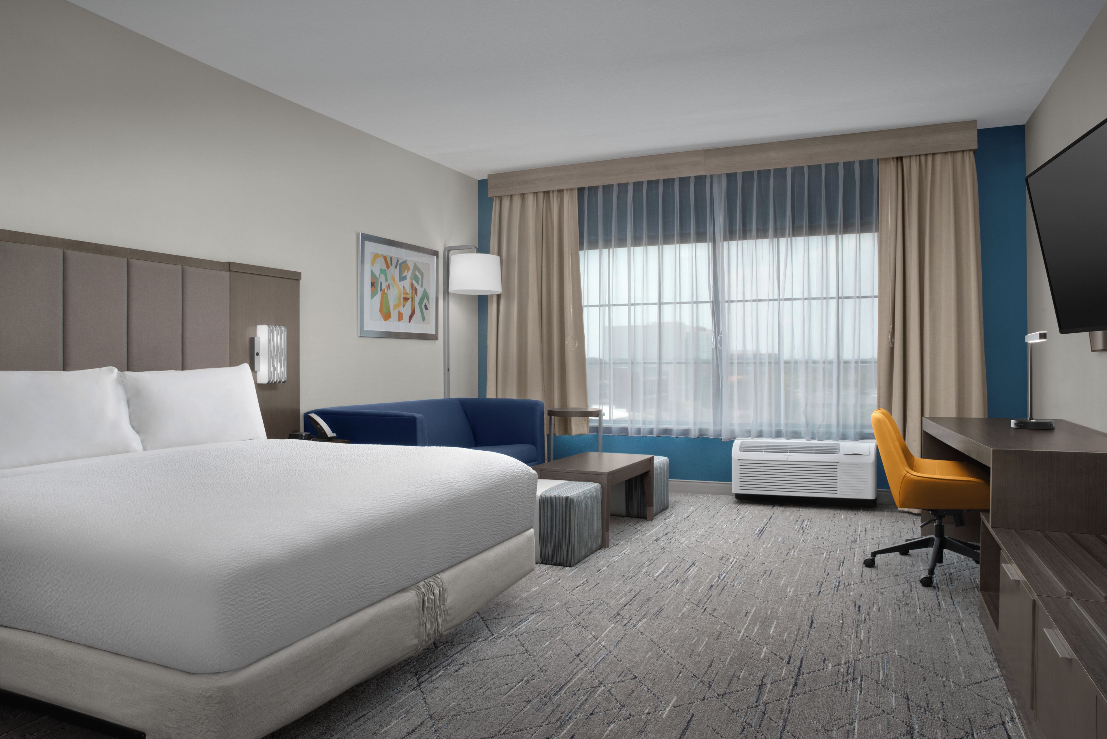 A hotel room with a bed, a desk, a chair, a sofa, a lamp, and a window with curtains.