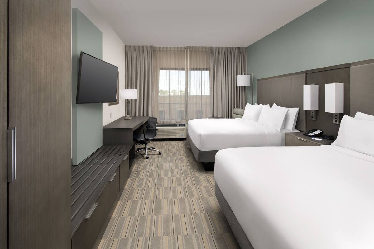 A hotel room with two beds, a desk, a flat-screen TV, and a window with curtains.