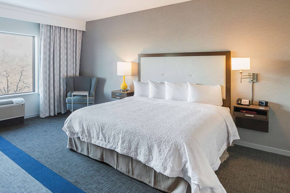 A hotel room with a large bed, two bedside tables with lamps, a chair, and a window with curtains.