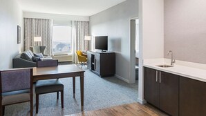A modern hotel room with a dining area, a TV, a sofa, and a kitchen area.