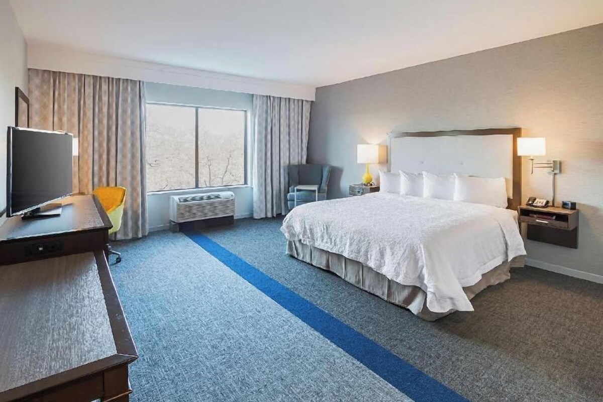 A hotel room with a large bed, a desk with a television, a chair, and a window with curtains.
