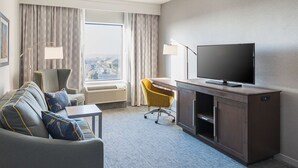 A hotel room with a grey sofa, yellow chair, TV, and a desk with a lamp.