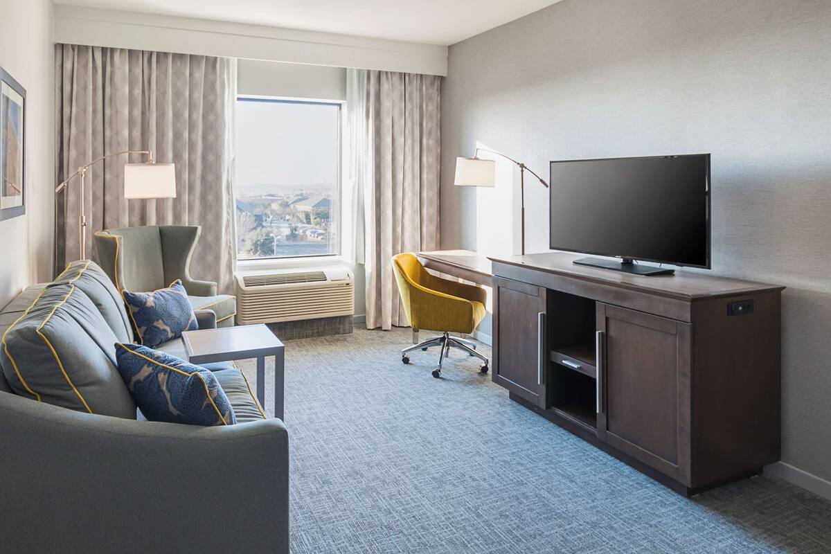 A hotel room with a grey sofa, yellow chair, TV, and a desk with a lamp.