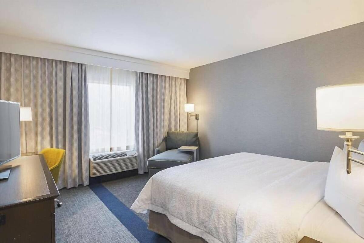 A hotel room with a bed, a desk, a chair, a lamp, and a window with curtains.