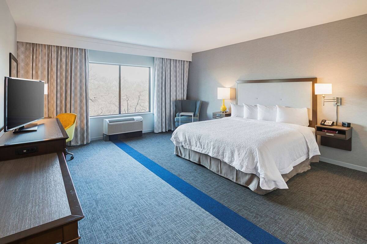 A hotel room with a bed, a desk with a TV, a chair, a bedside table with a lamp, and a window with curtains.