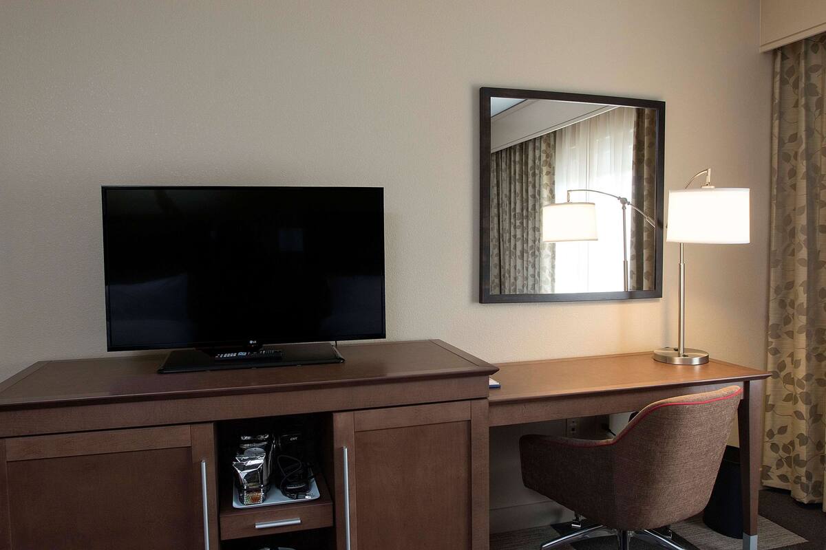 A hotel room with a flat-screen TV on a wooden desk, a mirror, a desk lamp, a chair, and a wardrobe with a shoe rack.