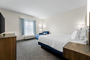A hotel room with a bed, a desk, a chair, a window with curtains, a standing lamp, and a radiator.