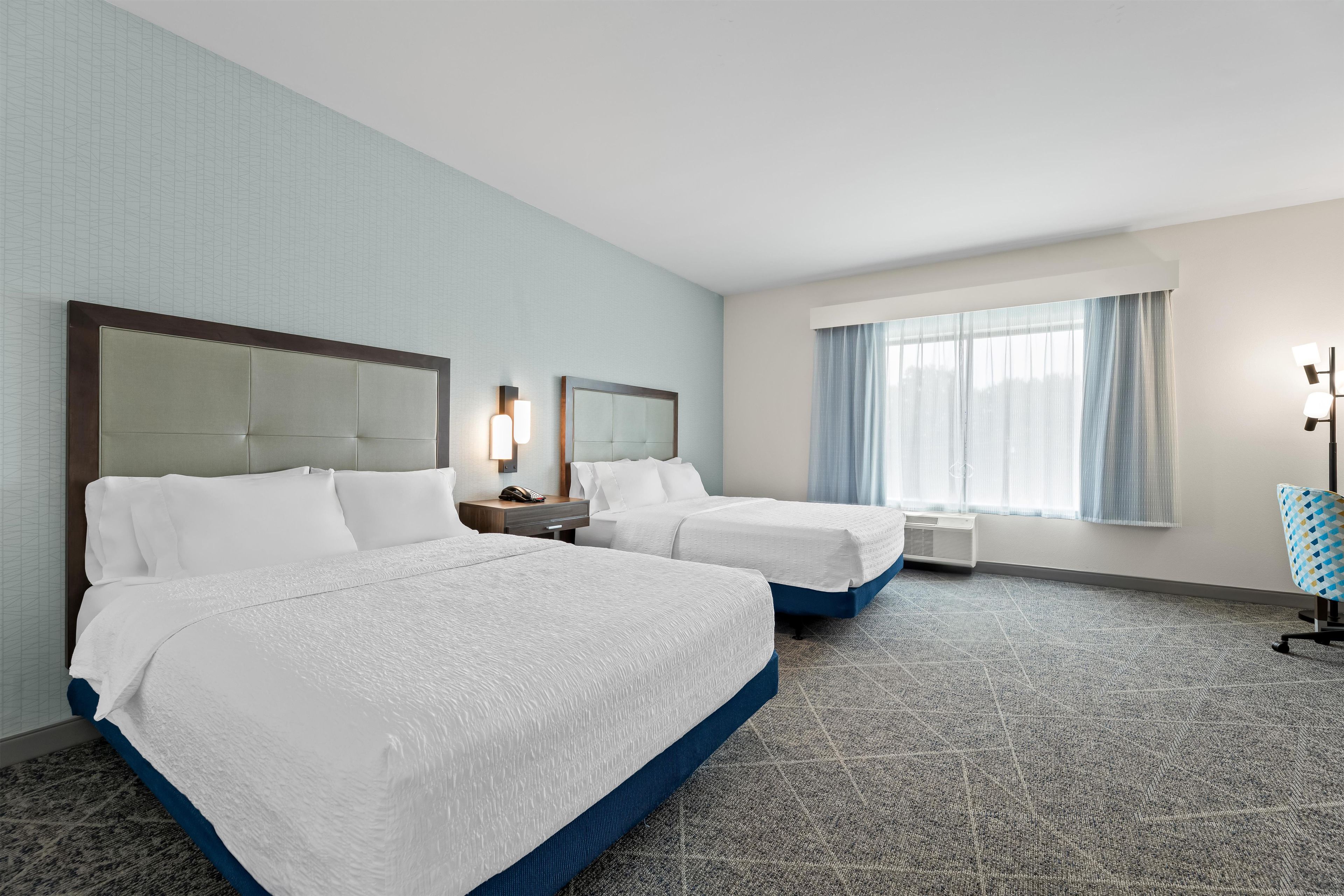Ein Hotelzimmer mit zwei Betten, einem Nachttisch, einer Lampe, einem Fenster mit Vorhängen und einem Teppichboden.
