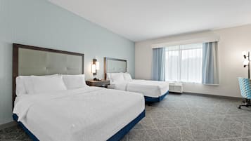 Habitación de hotel con dos camas, una mesita de noche, una lámpara, una ventana con cortinas y suelo alfombrado.