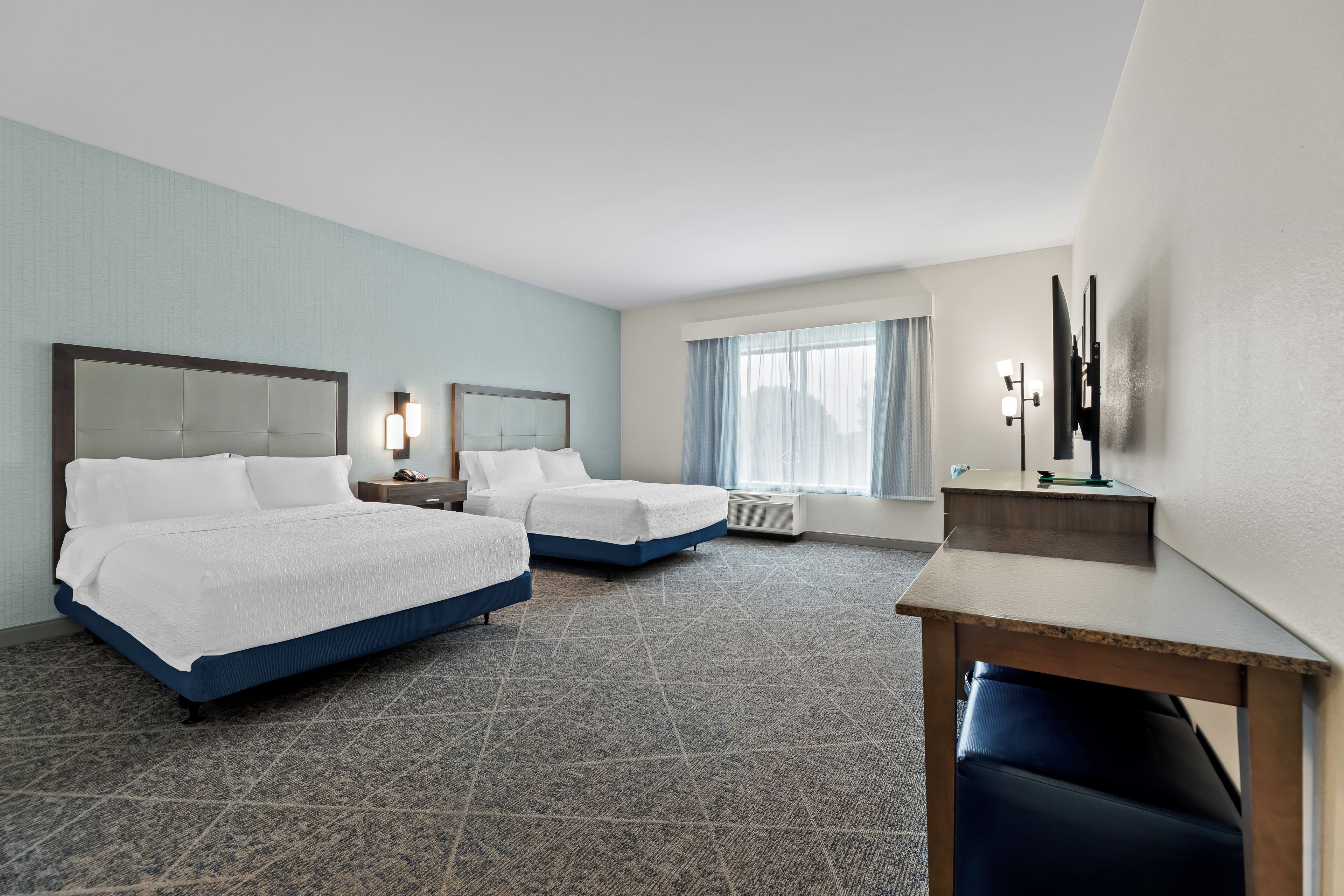 A hotel room with two beds, a desk, a chair, a TV, and a window with curtains.