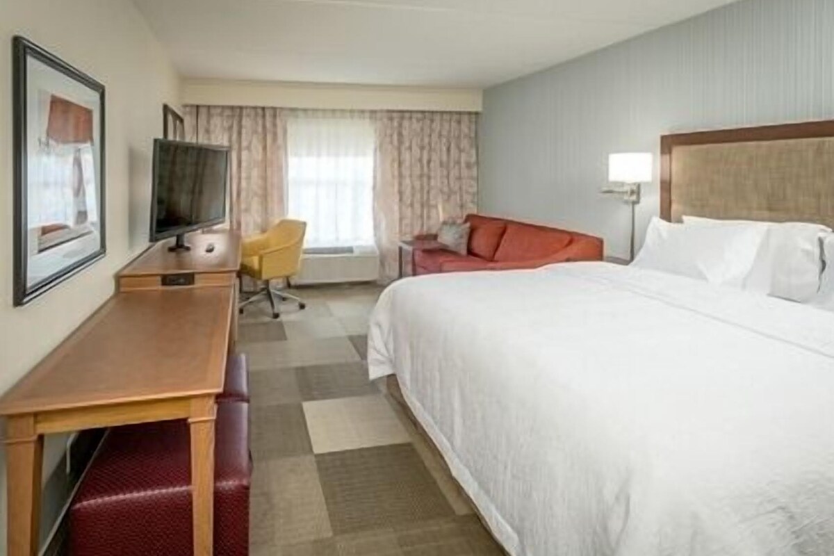 A hotel room with a bed, a desk with a chair, a television, and a window with curtains.