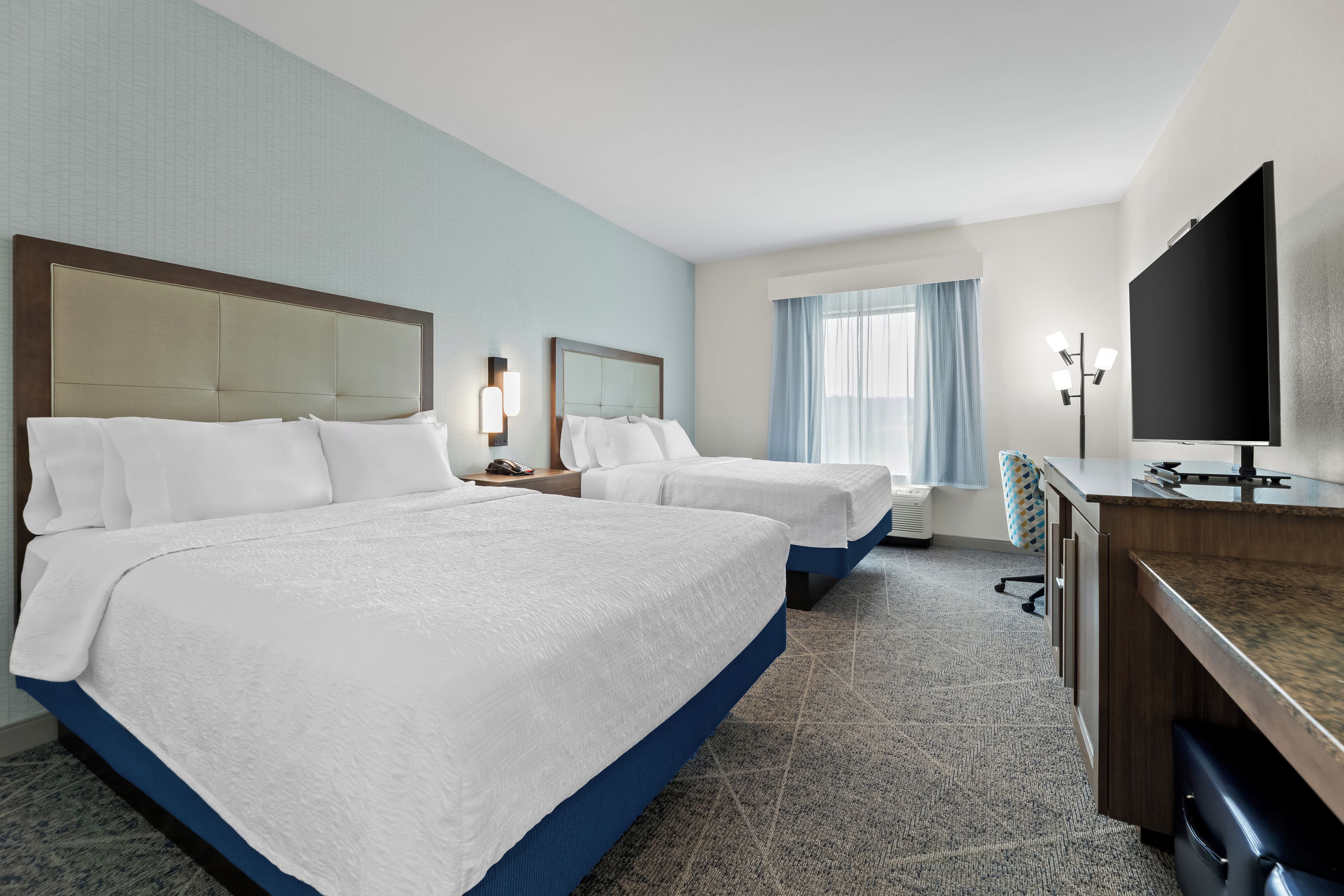 Une chambre d’hôtel avec deux lits, un bureau doté d’un téléviseur à écran plat, une lampe de bureau et une fenêtre avec des rideaux.
