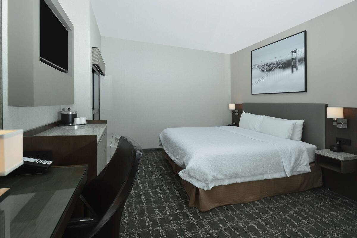A hotel room with a bed, a desk with a keyboard, a chair, a lamp, and a framed picture on the wall.
