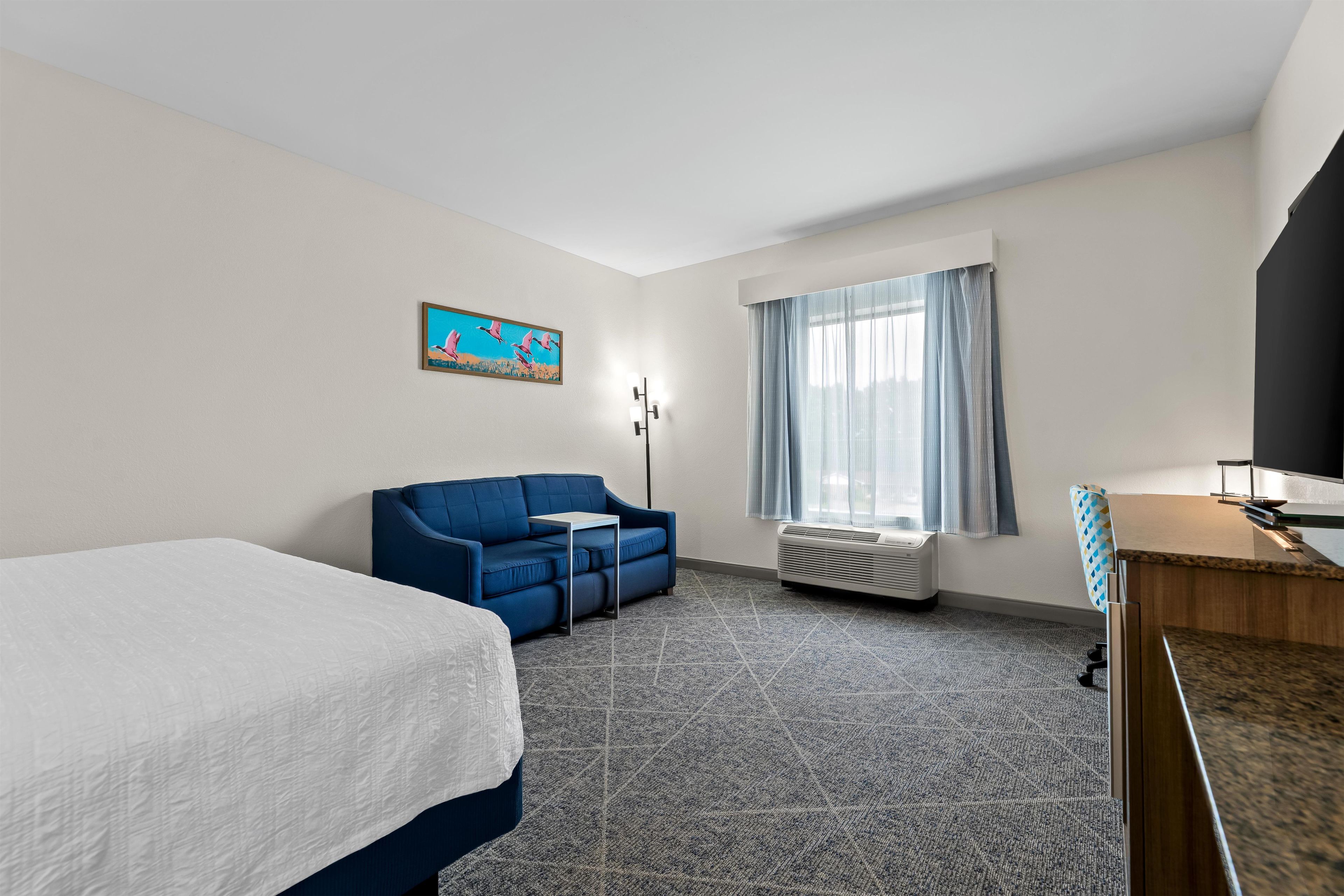 Una habitación de hotel con cama, sofá azul, una mesita, televisión y ventana con cortinas.