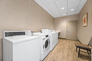 A laundry room with a commercial washer, a front-loading washing machine, and a drying machine. There is a bench and a door leading to another room.