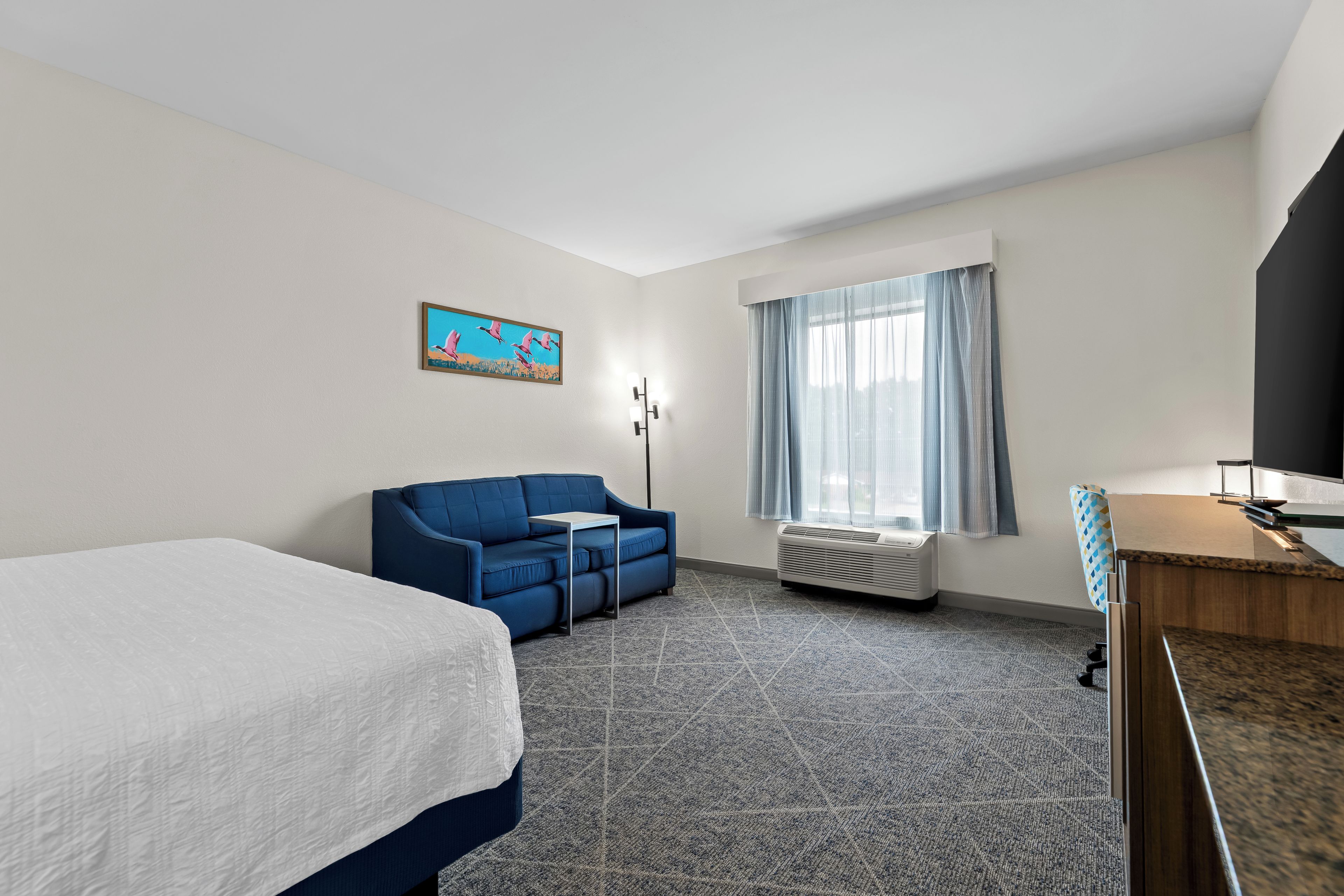 Ein Hotelzimmer mit einem Bett, einem blauen Sofa, einem kleinen Tisch, einem Fernseher und einem Fenster mit Vorhängen.