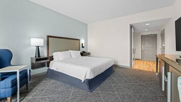 Habitación de hotel con cama, escritorio, silla, mesita de noche, lámpara y puerta que da a otra habitación.