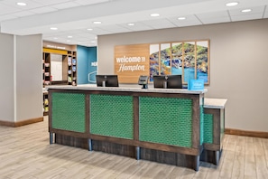 A hotel reception desk with a green-tiled backdrop, a large welcome sign, and a view of a forest scene.