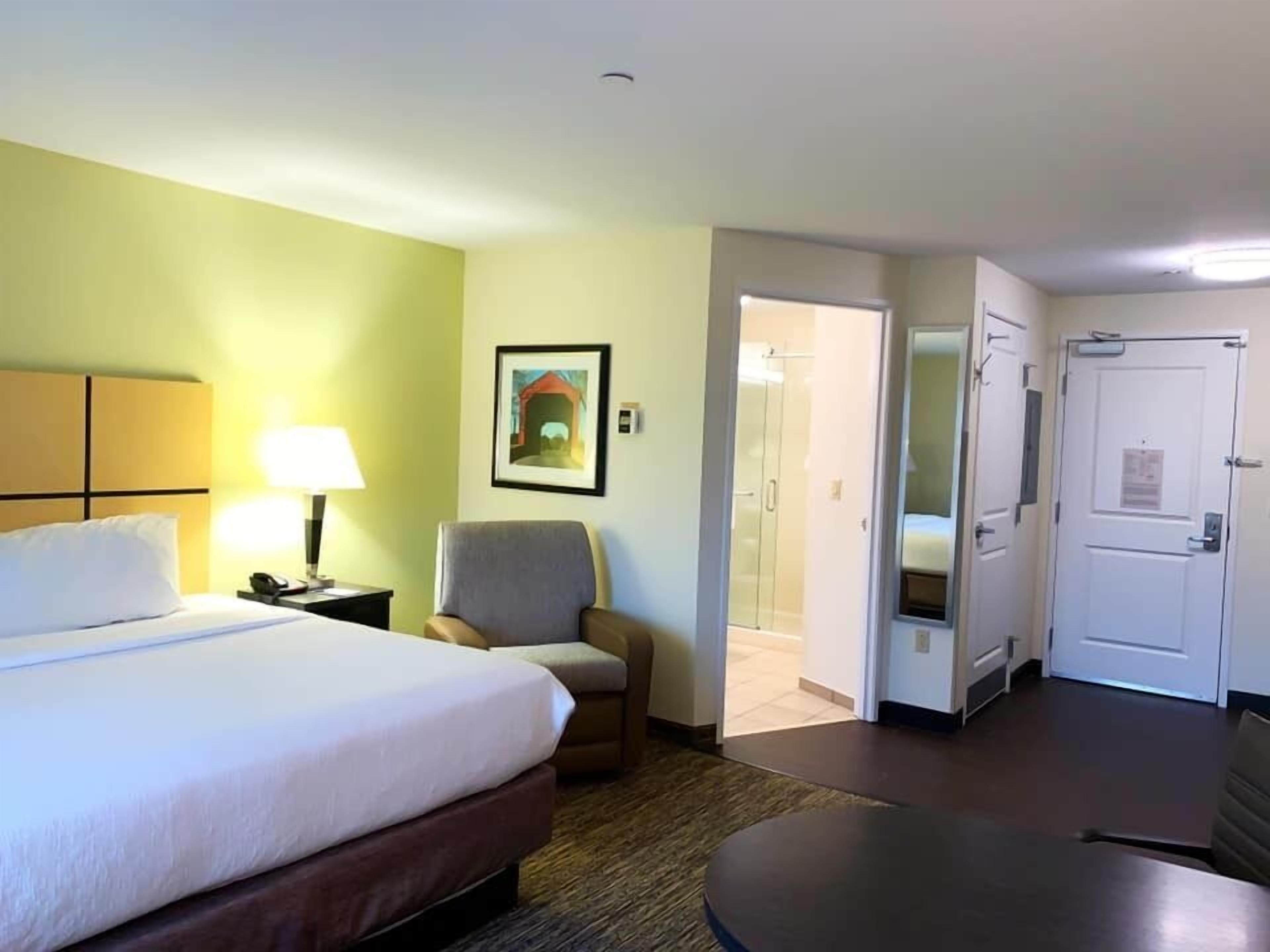 A hotel room with a bed, a chair, a desk, a lamp, a picture on the wall, and a door leading to a bathroom.
