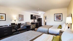 Quarto de hotel com duas camas, uma escrivaninha, uma cadeira, uma mesinha, uma área de cozinha e obras de arte nas paredes.