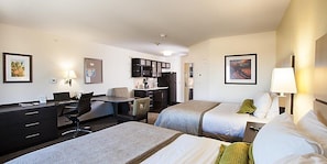 A hotel room with two beds, a desk, a chair, a small table, a kitchen area, and artwork on the walls. - Candlewood Suites Belle Vernon by IHG (Belle Vernon)