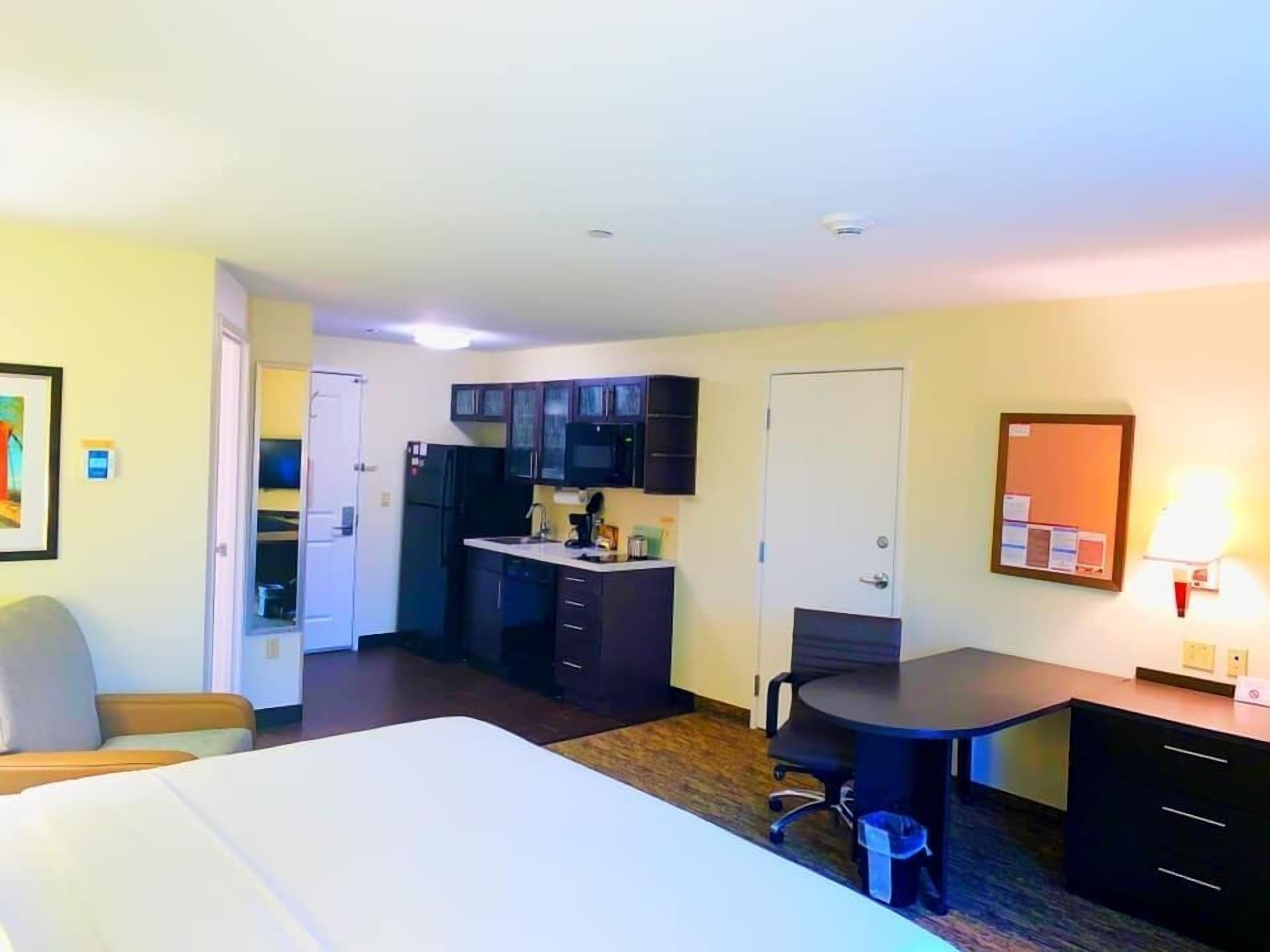 Una habitación de hotel con cama, escritorio, silla, kitchenette pequeño y puerta.