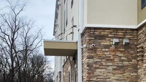 Een smal betonnen pad, begrensd door een stenen muur en een metalen hekwerk, leidt naar een gebouw met een plat dak en meerdere ramen.
