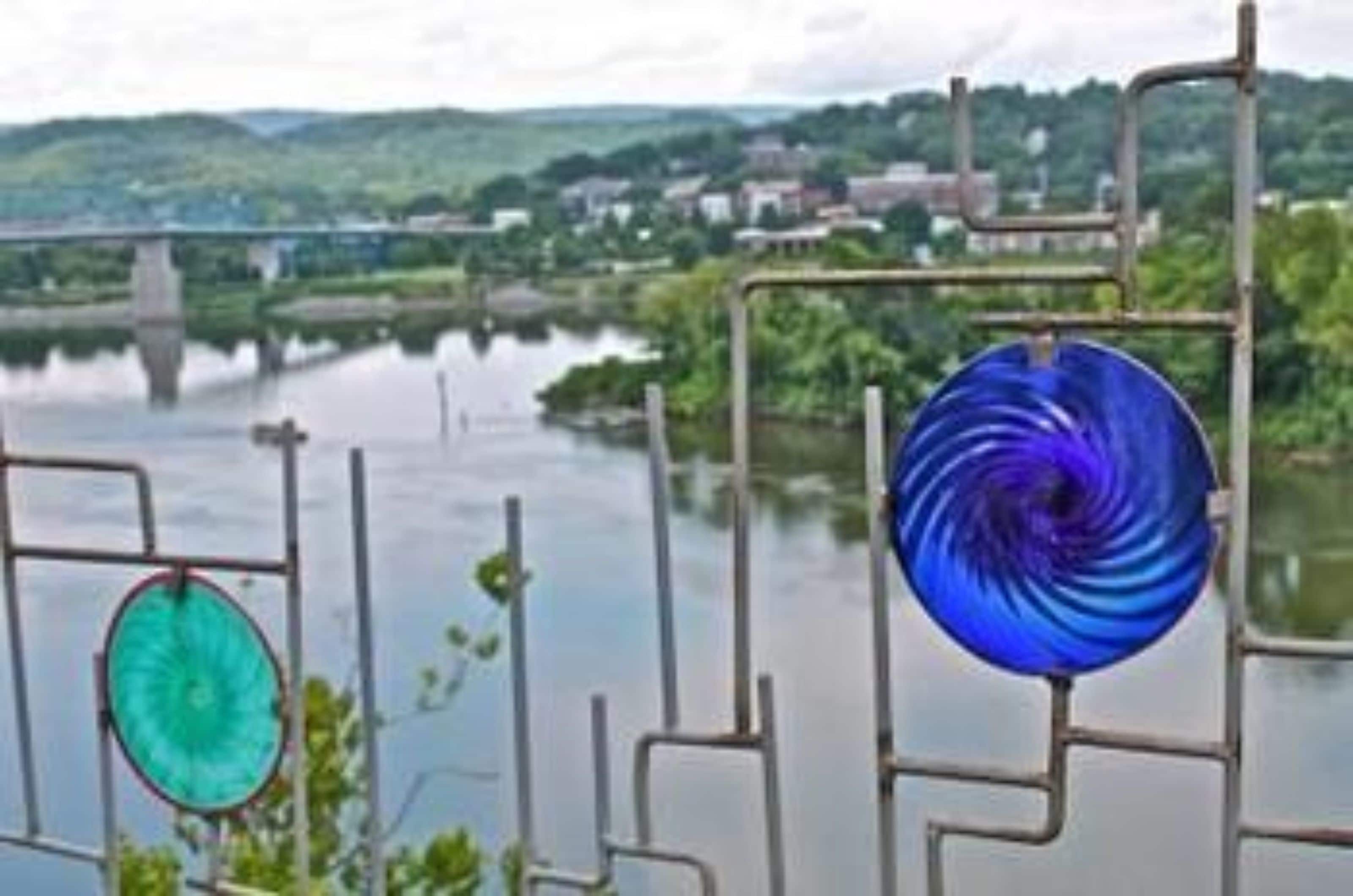 A metal railing with a blue and green circular object hanging from it, overlooking a river with a bridge and greenery in the background.