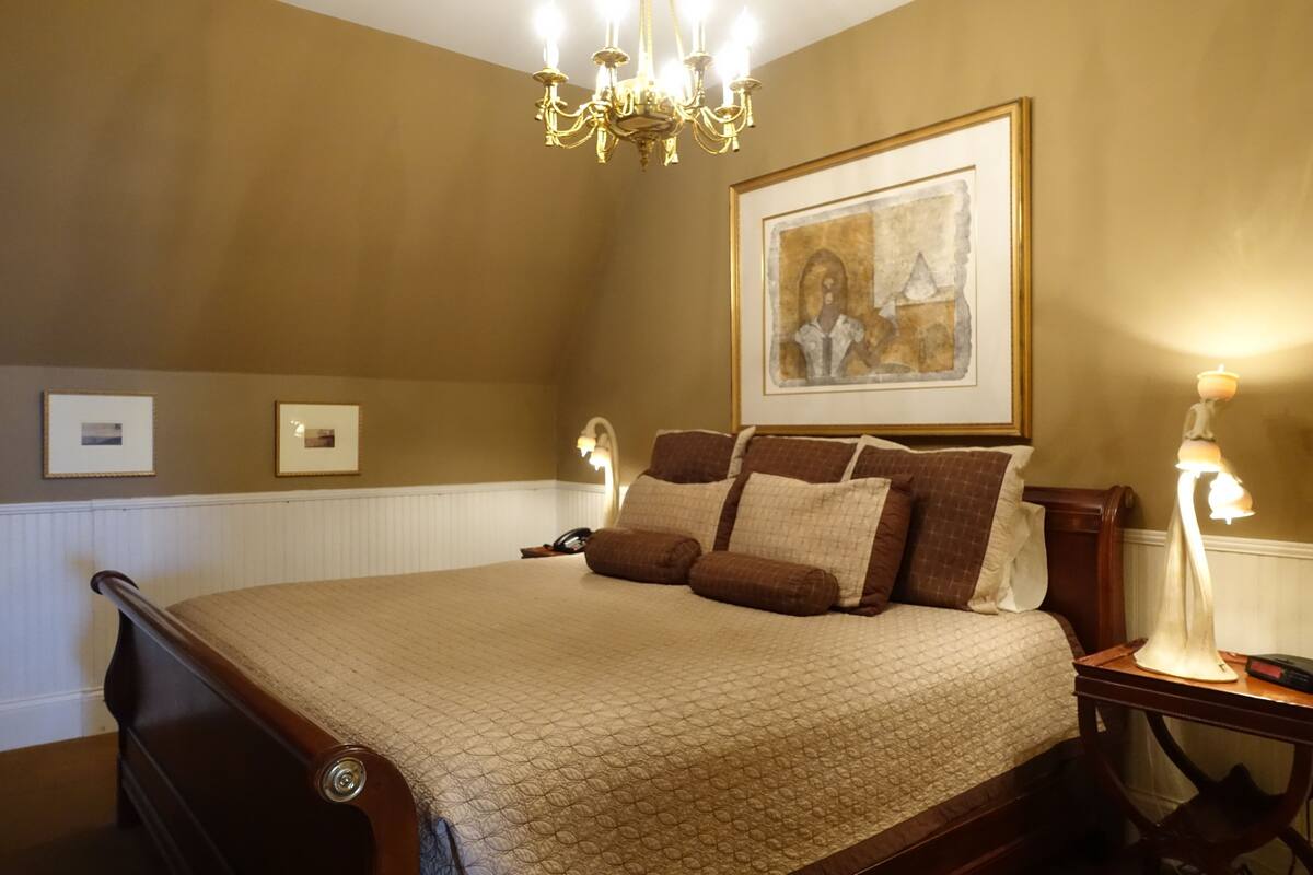 A bedroom with a large bed, a chandelier, a framed artwork on the wall, and a bedside table with a lamp.
