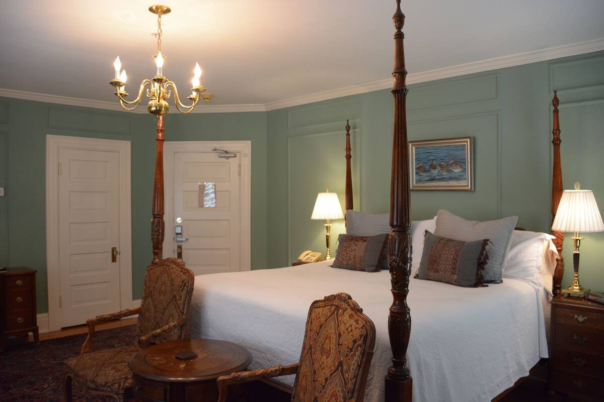 A four-poster bed with a white bedspread, two bedside tables with lamps, a chair, and a round table.