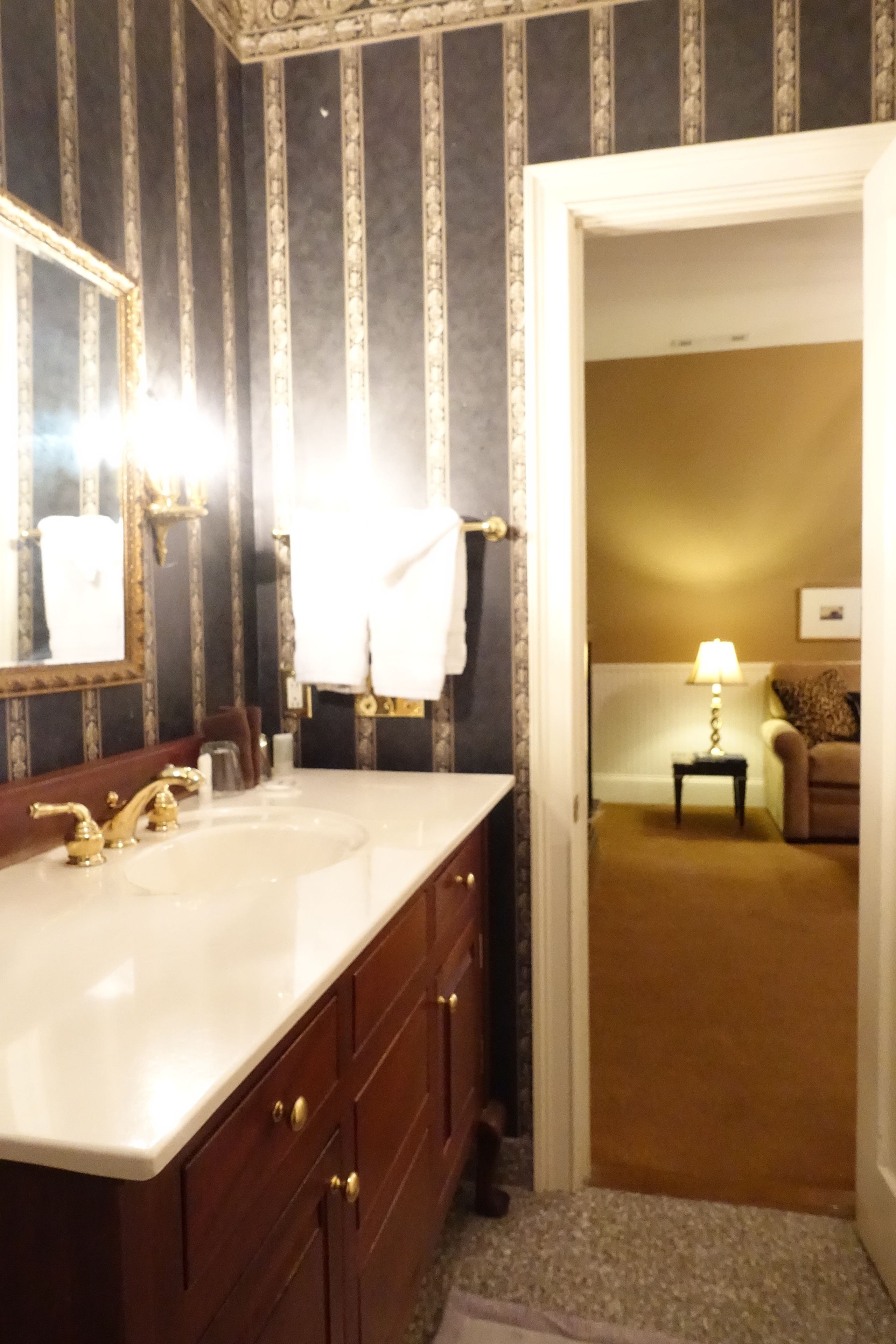 A bathroom with a large mirror, a white sink, and a vanity with wooden cabinets. There is a towel hanging on the wall and a small table with a lamp in the adjacent room.