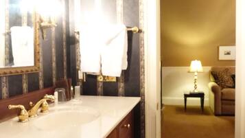 A bathroom with a large mirror, a white sink, and a vanity with wooden cabinets. There is a towel hanging on the wall and a small table with a lamp in the adjacent room.