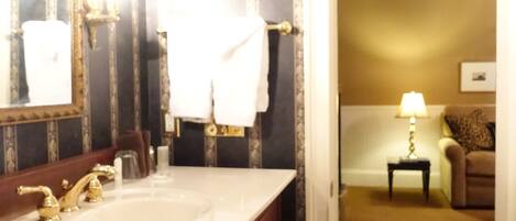 Une salle de bain avec un grand miroir, un lavabo blanc et un meuble-lavabo avec des placards en bois. Une serviette est accrochée au mur et, dans la pièce adjacente, on trouve une petite table avec une lampe.