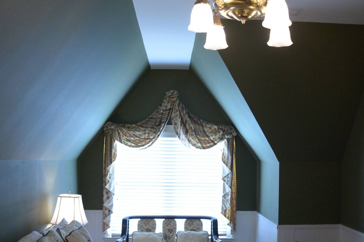 A bedroom with a sloped ceiling, a chandelier, a bed with a floral bedspread, and a window with curtains.