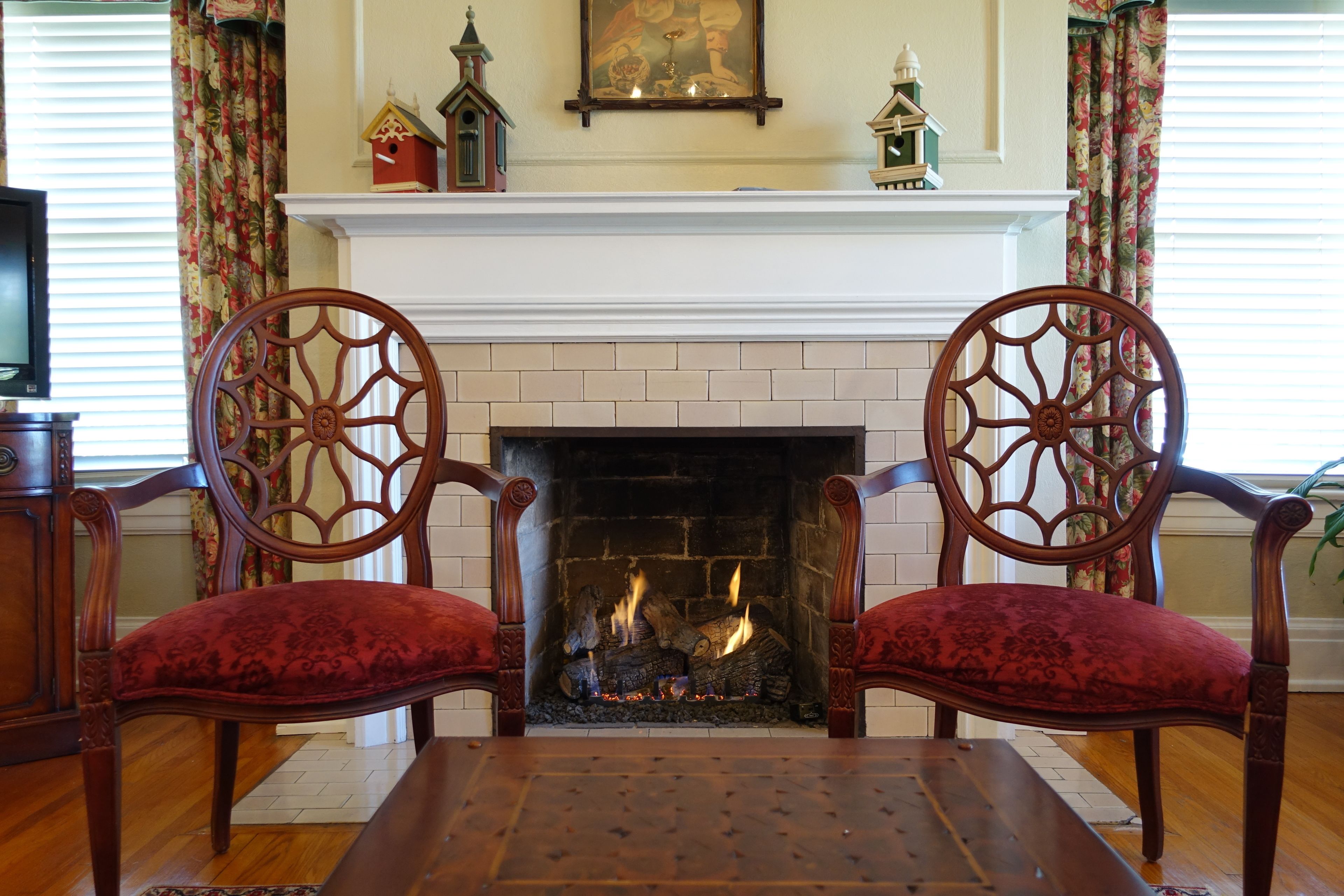 A fireplace with two ornate chairs facing it, a mantelpiece with decorative items, and a television on the left.
