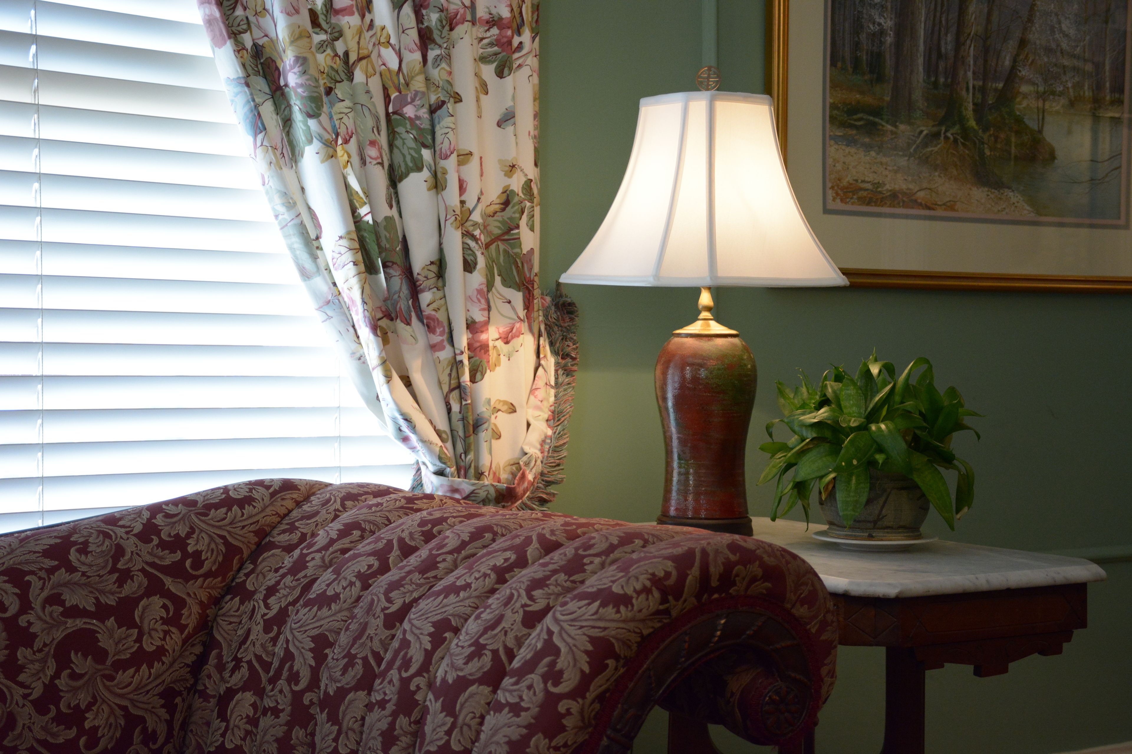 A bedroom with a bed, a lamp, a plant, and a painting on the wall.