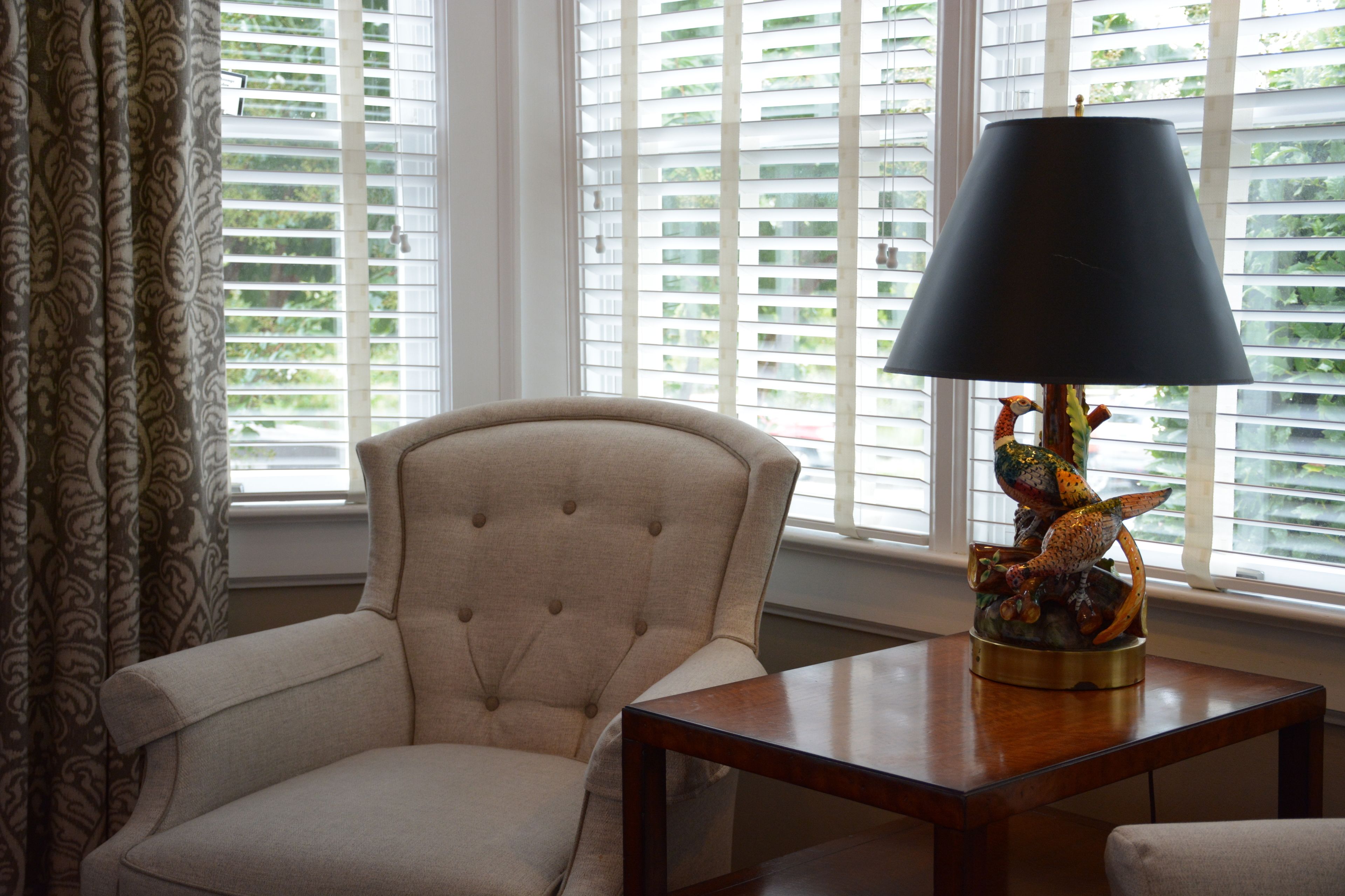 A room with a tufted armchair, a wooden table with a lamp, and a window with blinds.