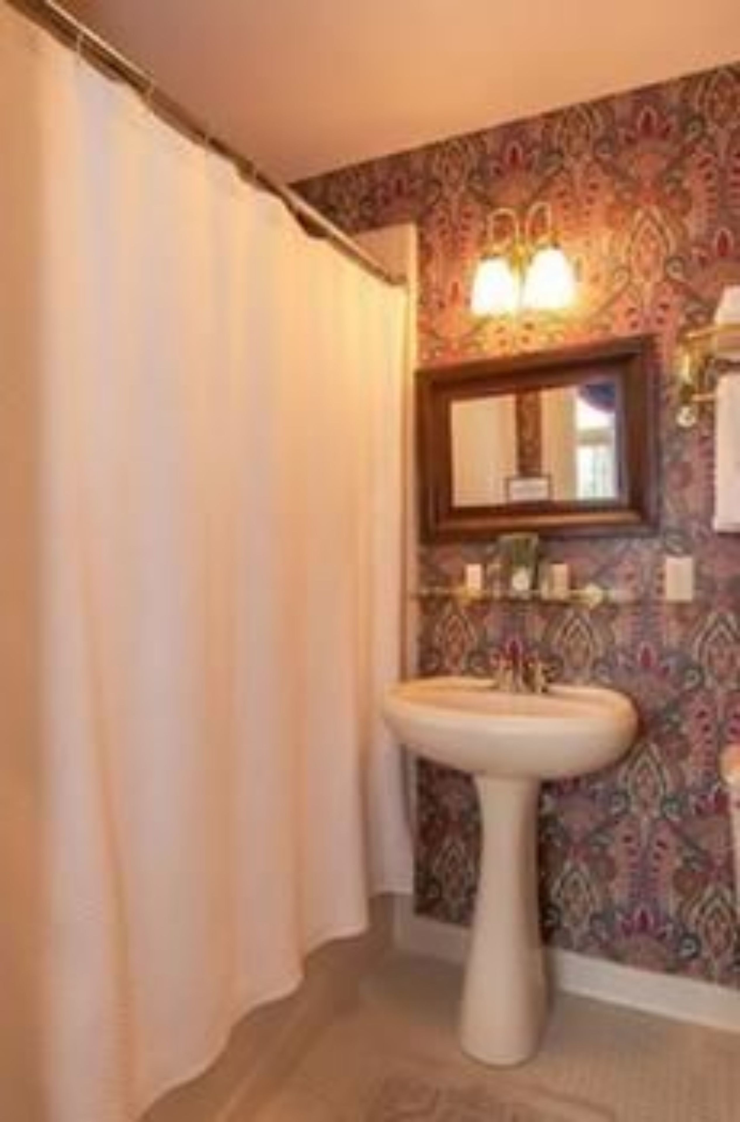 A bathroom with a pedestal sink, a mirror, and patterned wallpaper.