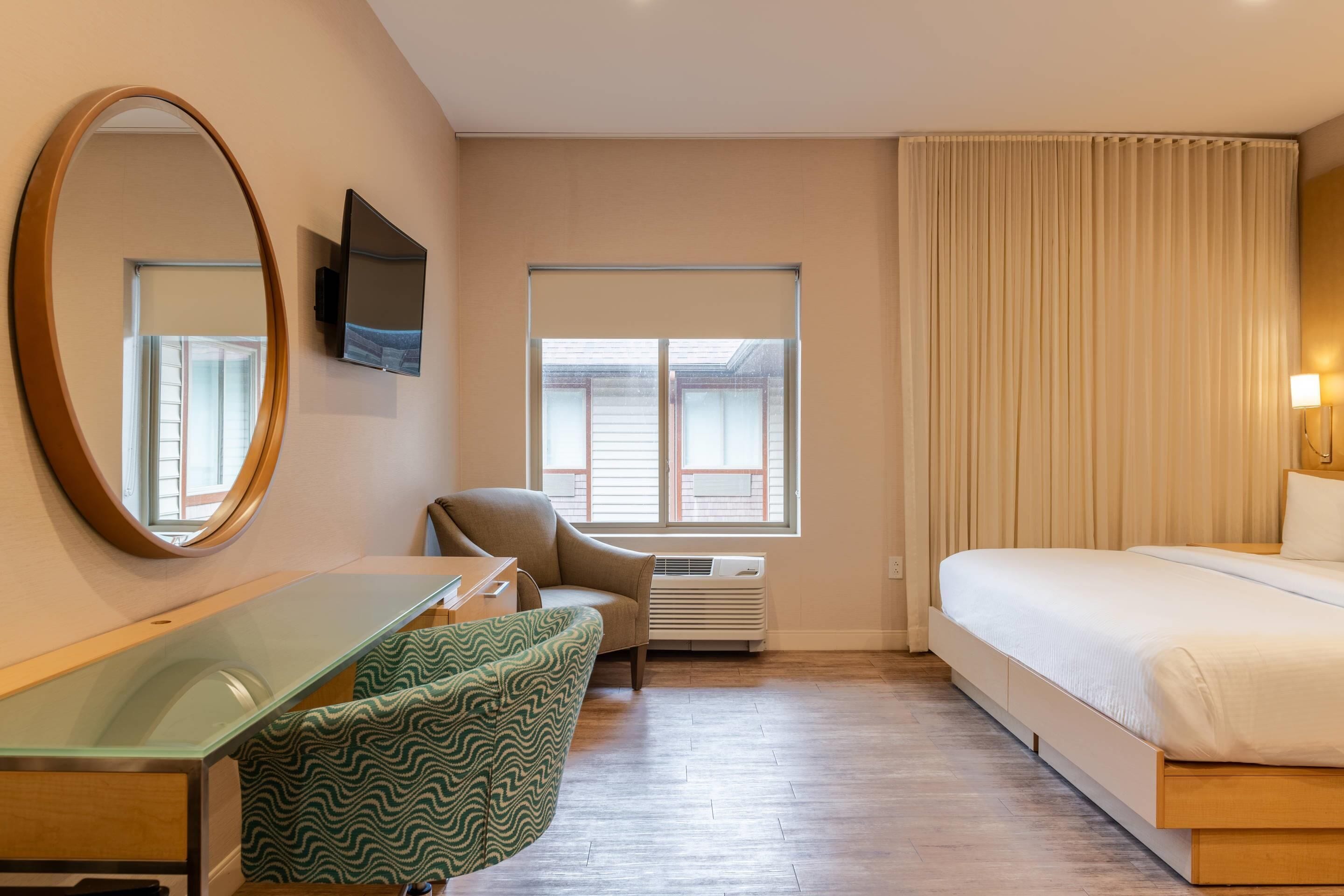 A hotel room with a bed, a desk, a chair, a large mirror, a flat-screen TV, a window with curtains, and an air conditioning unit.