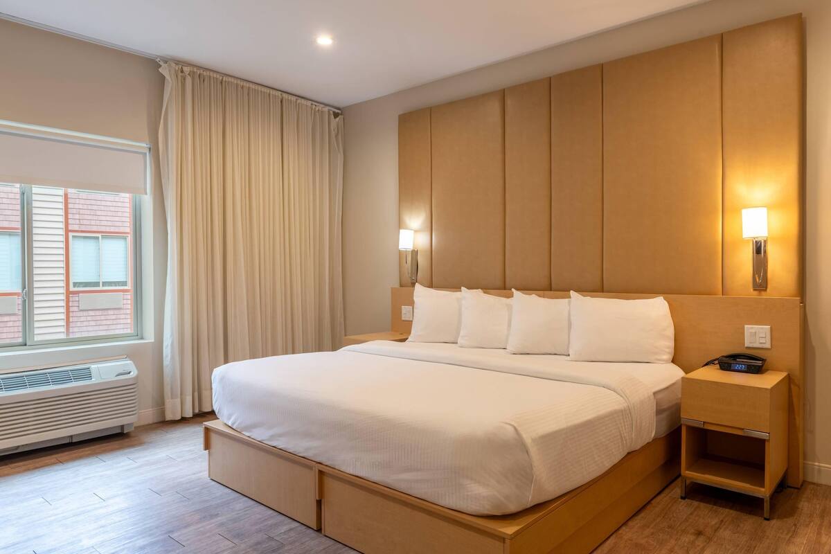 A hotel room with a large bed, a bedside table with a lamp, a window with curtains, and a wall-mounted heater.