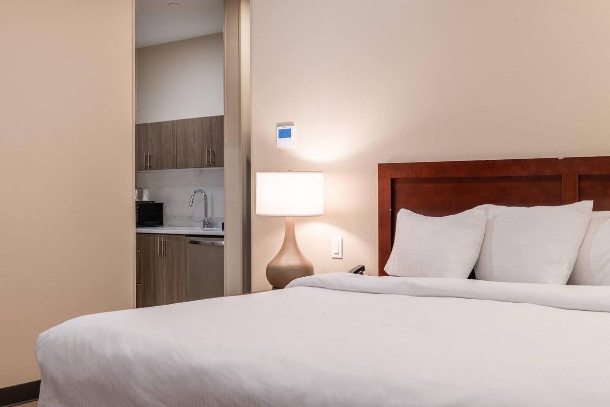 A hotel room with a bed, a nightstand, a lamp, a wall-mounted thermostat, and a view into the kitchen area.