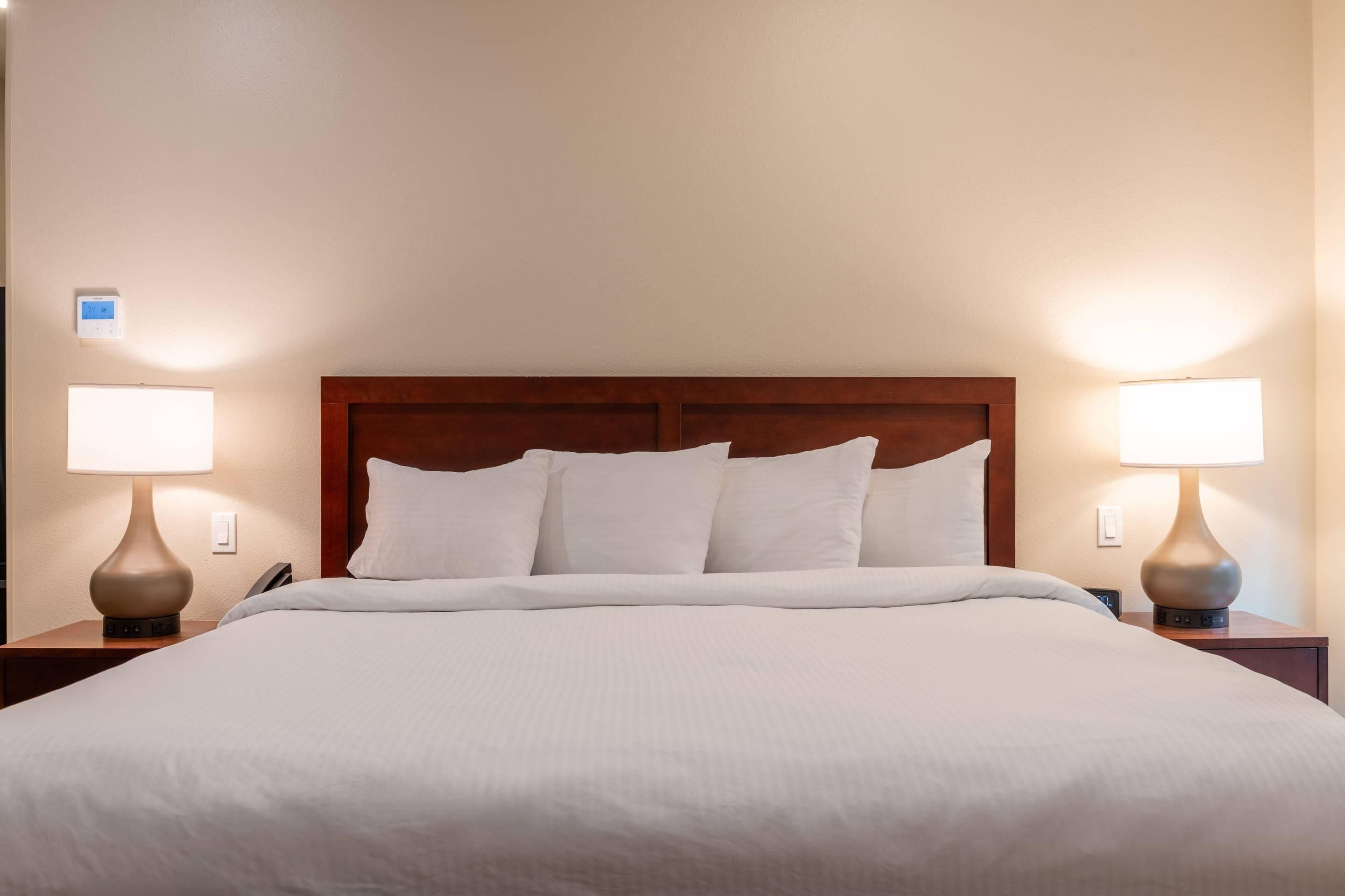 A neatly made bed with a wooden headboard, two bedside lamps, and a wall-mounted thermostat.